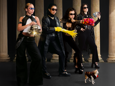 Four Latinx women pose in sunglasses looking mysterious holding a sparkly cloche, a yellow rubber glove, a red feather duster and a handful of passports. A chihuahua stands in a coat in front of them. 