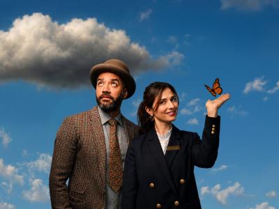 One Mixed race man in a brown tweed jacket and brown bowler hat looks up to a grey cloud in the sky. Next to him, a woman in a navy jacket with gold buttons looks into the camera as an orange butterfly lands on her hand. 