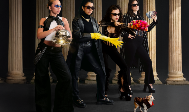 Four Latinx women pose in sunglasses looking mysterious holding a sparkly cloche, a yellow rubber glove, a red feather duster and a handful of passports. A chihuahua stands in a coat in front of them. 